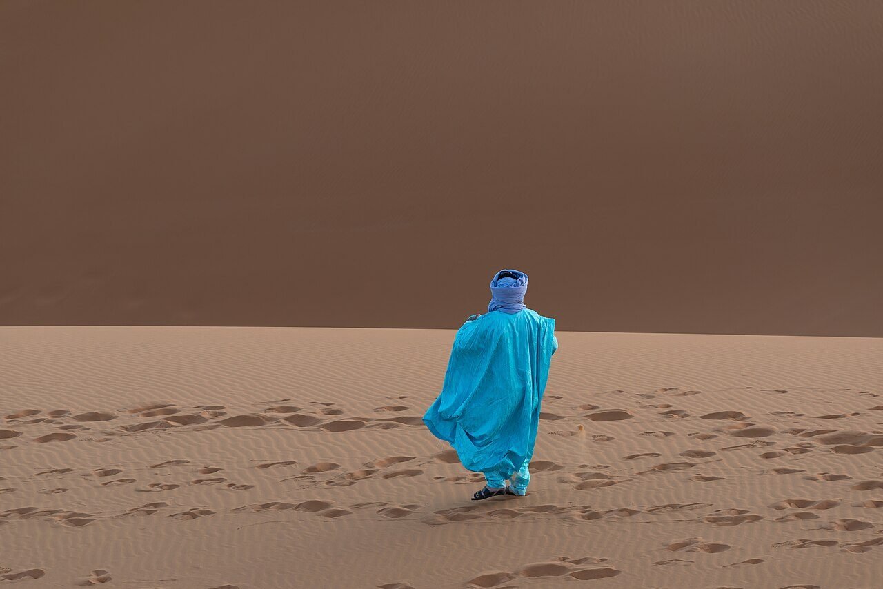 Traveller in a blue desert scarf and long sleeves sitting on a sand dune in the Moroccan Sahara with a small backpack
